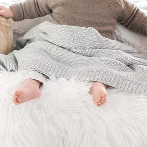 Könnte beinhalten: Die Füße eines Babys sind unter einer weichen, grauen Strickdecke sichtbar. Die Decke liegt auf einer weißen, flauschigen Oberfläche.