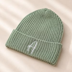 以下が含まれることがあります： 折り返しのあるセージグリーンのニット帽で、前面には白い刺繍で文字「A」が施されています。帽子全体が厚みのあるリブ編みのテクスチャーで構成されており、一貫した外観に仕上がっています。