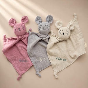 以下が含まれることがあります： 刺繍された顔を持つぬいぐるみのネズミの頭が付いた、3枚の柔らかいモスリン製セキュリティブランケット。ブランケットはピンク、グレー、クリーム色で、それぞれ結び目のある角と、Adelia、Lilly、Mateaという刺繍された名前が生地に入っています。