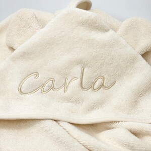 Puede incluir: Una toalla con capucha de color crema con una capucha en forma de oso y el nombre "Carla" bordado en color crema.