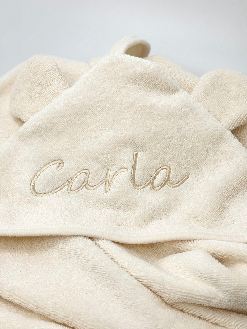 Puede incluir: Una toalla con capucha de color crema con una capucha en forma de oso y el nombre "Carla" bordado en color crema.