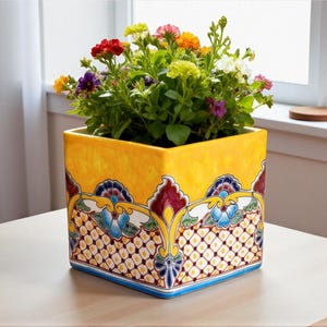 Può includere: Un vaso quadrato in ceramica gialla con un colorato motivo floreale e una varietà di fiori in piena fioritura. Il vaso ha un bordo blu e si trova su un tavolo colorato chiaro.