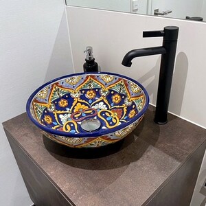 May include: A decorative, round ceramic sink with a vibrant, hand-painted floral design in blue, yellow, and red. The sink sits atop a dark brown cabinet, with a black faucet and soap dispenser nearby. The sink is a bathroom fixture.