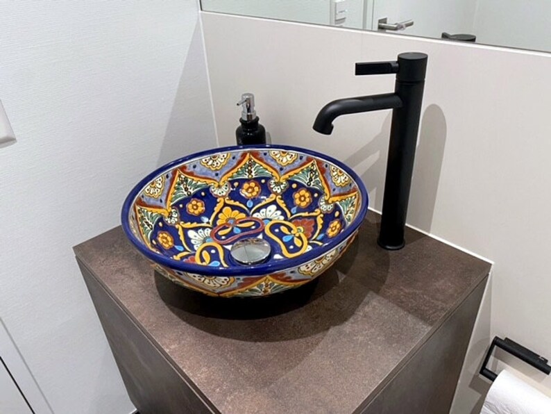 May include: A decorative, round ceramic sink with a vibrant, hand-painted floral design in blue, yellow, and red. The sink sits atop a dark brown cabinet, with a black faucet and soap dispenser nearby. The sink is a bathroom fixture.