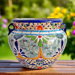 Puede incluir: Jardinera de cerámica pintada a mano con un vibrante diseño floral. La jardinera presenta un borde y asas azules, con intrincados patrones en verde, naranja y azul. Se encuentra sobre una superficie de madera, con un fondo borroso de flores coloridas.