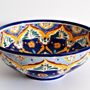 May include: A ceramic bowl with a vibrant, hand-painted design. The bowl features a deep blue rim and base, with a light blue interior. The interior is decorated with a repeating pattern of yellow, red, and green floral motifs. The bowl is likely 12 inches in diameter.