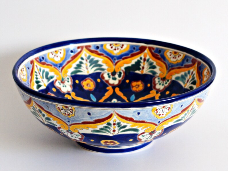 May include: A ceramic bowl with a vibrant, hand-painted design. The bowl features a deep blue rim and base, with a light blue interior. The interior is decorated with a repeating pattern of yellow, red, and green floral motifs. The bowl is likely 12 inches in diameter.