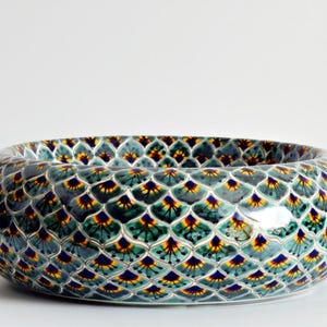 May include: Oval ceramic bowl with a repeating pattern of teal, yellow, and navy blue shapes. The bowl has a glossy finish and a wide, shallow design. The pattern resembles overlapping scales or feathers. The bowl is likely used for decorative purposes.