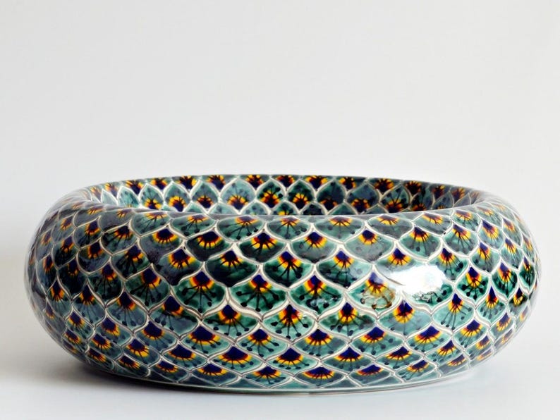 May include: Oval ceramic bowl with a repeating pattern of teal, yellow, and navy blue shapes. The bowl has a glossy finish and a wide, shallow design. The pattern resembles overlapping scales or feathers. The bowl is likely used for decorative purposes.