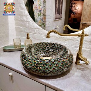 May include: A decorative ceramic sink with a green, yellow, and blue peacock feather pattern. The sink is round and sits on a gray countertop. A gold faucet and other bathroom accessories are visible in the background.
