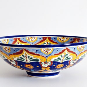 May include: A large, round ceramic bowl with a vibrant, hand-painted design. The bowl features a deep blue rim and base, with a colorful pattern of yellow, red, and green floral motifs on a light blue background. The bowl is approximately 12 inches in diameter.