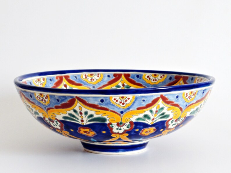 May include: A large, round ceramic bowl with a vibrant, hand-painted design. The bowl features a deep blue rim and base, with a colorful pattern of yellow, red, and green floral motifs on a light blue background. The bowl is approximately 12 inches in diameter.