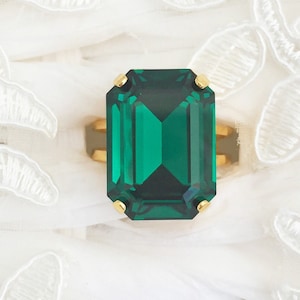 May include: A gold ring with a large, emerald-cut, deep green gemstone. The ring has a simple setting with four small gold prongs. The background is a soft, white fabric with delicate lace detailing. The ring's design is elegant.