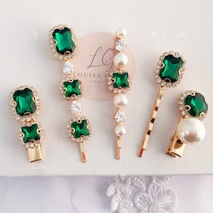 Puede incluir: Una variedad de accesorios para el cabello dorados, que incluyen horquillas y pasadores. Cada pieza presenta piedras preciosas rectangulares y cuadradas de color verde esmeralda, rodeadas de cristales claros más pequeños. Algunos diseños incorporan perlas de imitación.