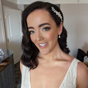 May include: A bride with dark hair styled in loose waves, wearing a white beaded gown and a pearl hairpiece. She is smiling and looking at the camera.