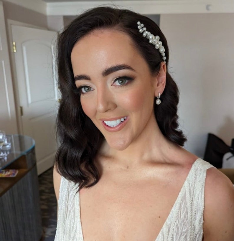 May include: A bride with dark hair styled in loose waves, wearing a white beaded gown and a pearl hairpiece. She is smiling and looking at the camera.