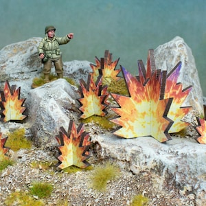 Puede incluir: Un conjunto de siete efectos de explosión en miniatura de cartón para juegos de guerra de mesa. Las explosiones están pintadas en tonos de amarillo, naranja y rojo, con un tono púrpura en los bordes. Las explosiones están dispuestas sobre una superficie rocosa.