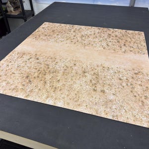May include: A square gaming mat featuring a printed desert landscape with a central dirt path, light brown sandy textures, and scattered patches of dry vegetation, designed for tabletop miniature wargaming and terrain displays.