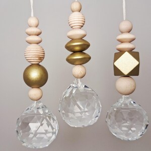 Könnte beinhalten: Drei hängende Zierornamente. Jedes Ornament hat eine klare Kristallkugel am unteren Ende, darüber Holzperlen und goldfarbene Akzente. Die Perlen haben verschiedene Formen, darunter Kugeln, Ovale und ein Quadrat.