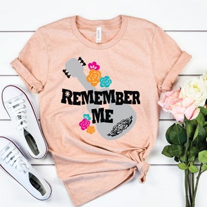 May include: A peach-colored t-shirt with a gray guitar silhouette and the words "Remember Me" in black. The guitar is decorated with colorful flowers and a skull.