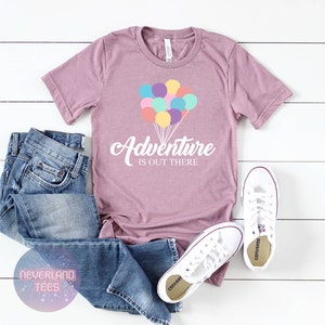 May include: A mauve t-shirt with a white graphic of colorful balloons and the text "Adventure is out there". The t-shirt is paired with blue jeans and white sneakers.