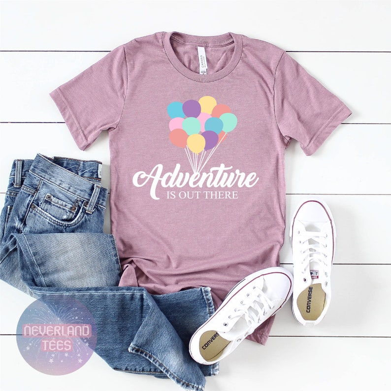May include: A mauve t-shirt with a white graphic of colorful balloons and the text "Adventure is out there". The t-shirt is paired with blue jeans and white sneakers.