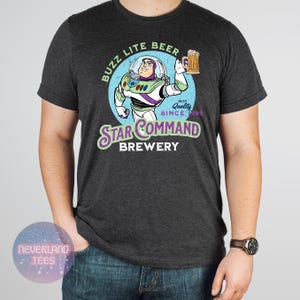 May include: Dark gray t-shirt featuring a cartoon image of Buzz Lightyear holding a beer mug, with the text "Buzz Lite Beer" and "Star Command Brewery". The design includes the words "Best Quality Since 1995".