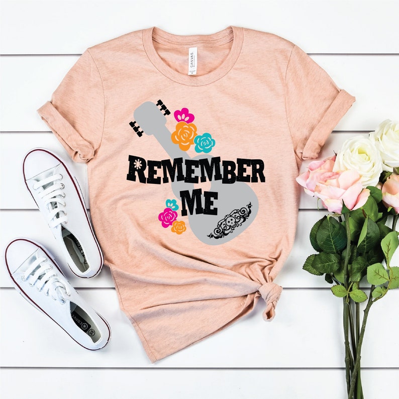May include: A peach-colored t-shirt with a gray guitar silhouette and the words "Remember Me" in black. The guitar is decorated with colorful flowers and a skull.