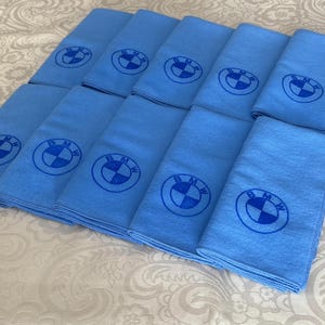 May include: A stack of light blue microfiber towels, each embroidered with the BMW logo in a darker shade of blue. The towels are neatly folded and arranged on a patterned surface.