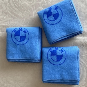 May include: Three light blue microfiber cleaning cloths with the BMW logo printed in dark blue. The BMW logo is a circle with the letters BMW and a four-quadrant design. The cloths are folded and stacked.