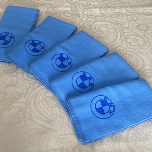 May include: Several light blue microfiber cloths with the BMW logo printed in dark blue. The cloths are arranged in a diagonal pattern on a patterned white surface. The BMW logo is a blue and white roundel.