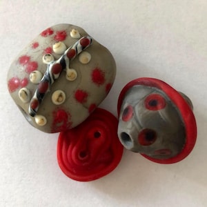 Puede incluir: Una colección de cuentas y botones de cerámica hechos a mano. Las cuentas son grises con manchas rojas y detalles blancos. Una cuenta tiene una raya en blanco y negro. También hay un botón rojo y un botón gris con borde rojo.