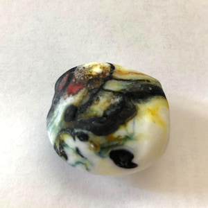 Puede incluir: Una piedra lisa y redondeada con un patrón de colores único. La piedra presenta una base blanca con remolinos negros, amarillos, rojos y verdes. La superficie de la piedra está pulida y parece natural.
