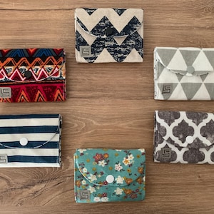 Könnte beinhalten: Eine Sammlung von sechs kleinen Stoffgeldbörsen mit verschiedenen Mustern, darunter Streifen, Chevron, Blumen und geometrische Designs. Jede Geldbörse hat einen Knopfverschluss und ein kleines quadratisches Logo. Auf einer Holzoberfläche angeordnet.