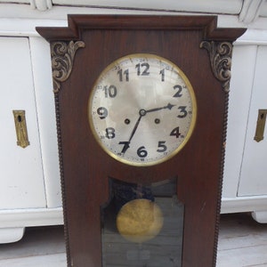 May include: Antique wooden wall clock with a round, white face and black Roman numeral markings. The clock has a decorative, ornate frame with gold accents.