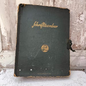 Op de afbeelding: Een vintage donkergroene ordner met het woord "Schriftenordner" in gouden letters. De ordner heeft een zwarte sluiting en een gouden embleem. De randen vertonen slijtage, wat op leeftijd duidt. De ordner ligt op een witte kanten doek.