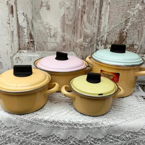 May include: Four vintage enamelware pots in varying sizes, each with a yellow body and a lid in pastel colors: yellow, pink, and light blue. The pots have black handles and lids, and are arranged on a white lace tablecloth.