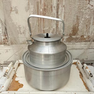 Op de afbeelding: Een vintage aluminium waterkoker met een handvat en een deksel, bovenop een grotere aluminium pot. De waterkoker en pot staan op een witte, verweerde houten dienblad met witte handvatten. De achtergrond is een verweerd wit oppervlak.