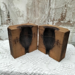 May include: Two halves of a vintage wooden mold, likely for glassblowing, with a dark interior and a light brown exterior. The mold is open, revealing the interior shape. The wood has visible grain and is secured with metal pins.