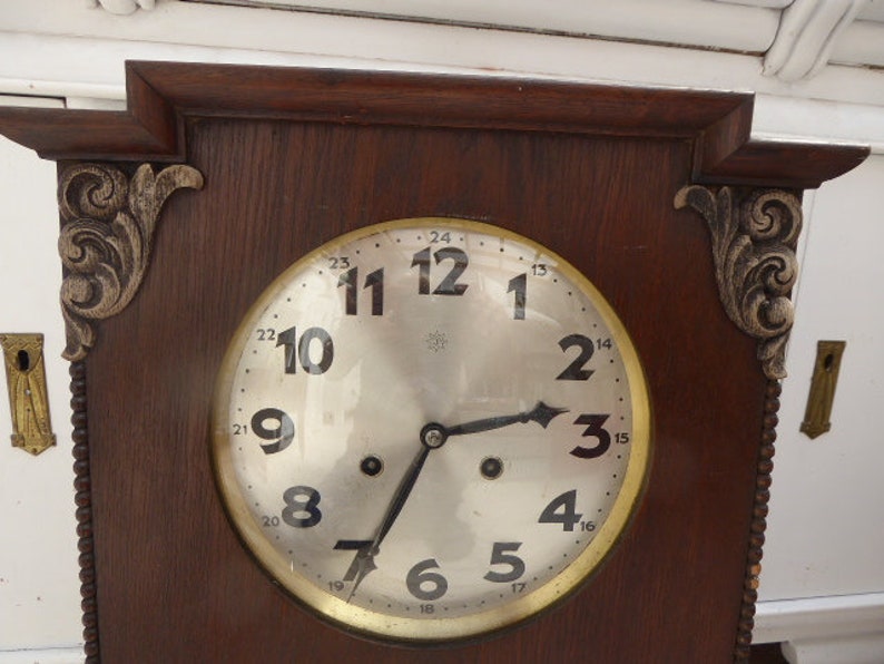 May include: A vintage wooden clock with a round, white face and black hands. The clock has a decorative, ornate design around the edge of the face and a dark wood frame.