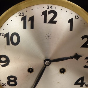 May include: A vintage clock face with black Roman numeral hour markers on a white background. The clock face is surrounded by a gold-colored metal frame with black markings for the minutes.