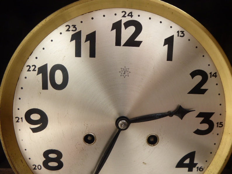 May include: A vintage clock face with black Roman numeral hour markers on a white background. The clock face is surrounded by a gold-colored metal frame with black markings for the minutes.