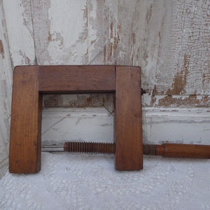 Könnte beinhalten: Eine alte Holzklammer mit Schraubmechanismus. Die Klammer ist aus dunkelbraunem Holz gefertigt und hat einen rechteckigen Rahmen. Die Schraube ist aus Metall mit einem Holzgriff. Der Hintergrund ist eine weiße, verwitterte Holztür.