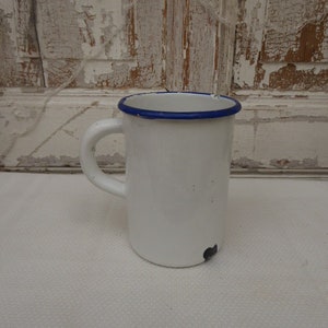 May include: White enamel mug with a blue rim. The mug has a handle and is sitting on a white surface.