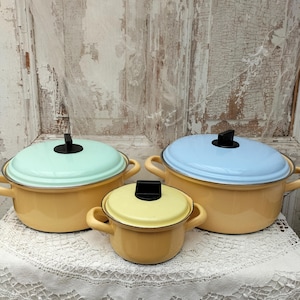 May include: Three vintage enamelware pots in varying sizes. The pots have yellow bodies with pastel green, blue, and yellow lids. Each pot has a black handle on the lid and two side handles. The pots are arranged on a white lace tablecloth.