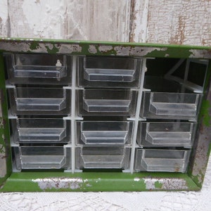 May include: A vintage green metal drawer organizer with 12 clear plastic drawers. The drawers are divided into two rows of six. The organizer has a distressed look with some rust and paint chips.