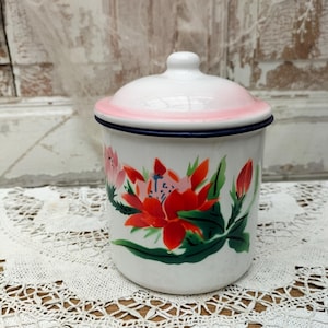 Könnte beinhalten: Ein weißer Emaille-Behälter mit passendem Deckel, der einen rosa Rand und eine dunkelblaue Kante aufweist. Der zylindrische Behälter ist mit einem lebendigen Blumenmuster aus roten und rosa Blüten sowie grünen Blättern auf der Vorderseite verziert.