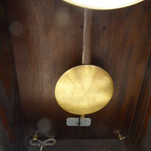 May include: A wooden clock face with a gold pendulum and a key wind mechanism. The clock face has black Roman numerals and a white background.