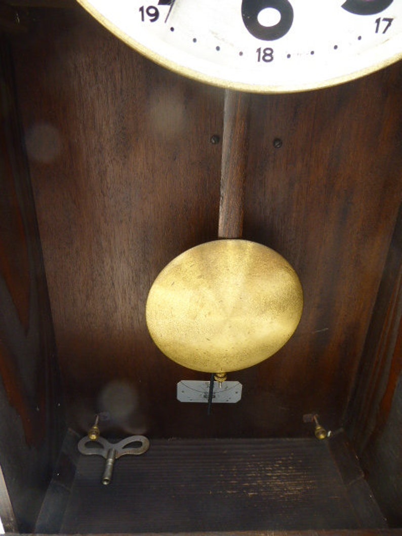 May include: A wooden clock face with a gold pendulum and a key wind mechanism. The clock face has black Roman numerals and a white background.