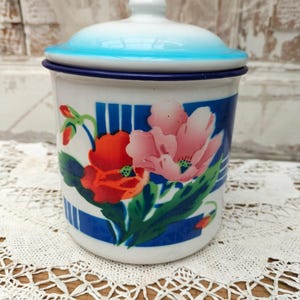 Könnte beinhalten: Ein weißer Emaille-Behälter mit Deckel und hellblauem Rand. Der Körper ist mit einem Blumenmuster aus roten und rosa Mohnblumen, grünen Blättern und vertikalen blauen Streifen verziert. Der Behälter hat eine zylindrische Form und eine glatte Oberfläche.